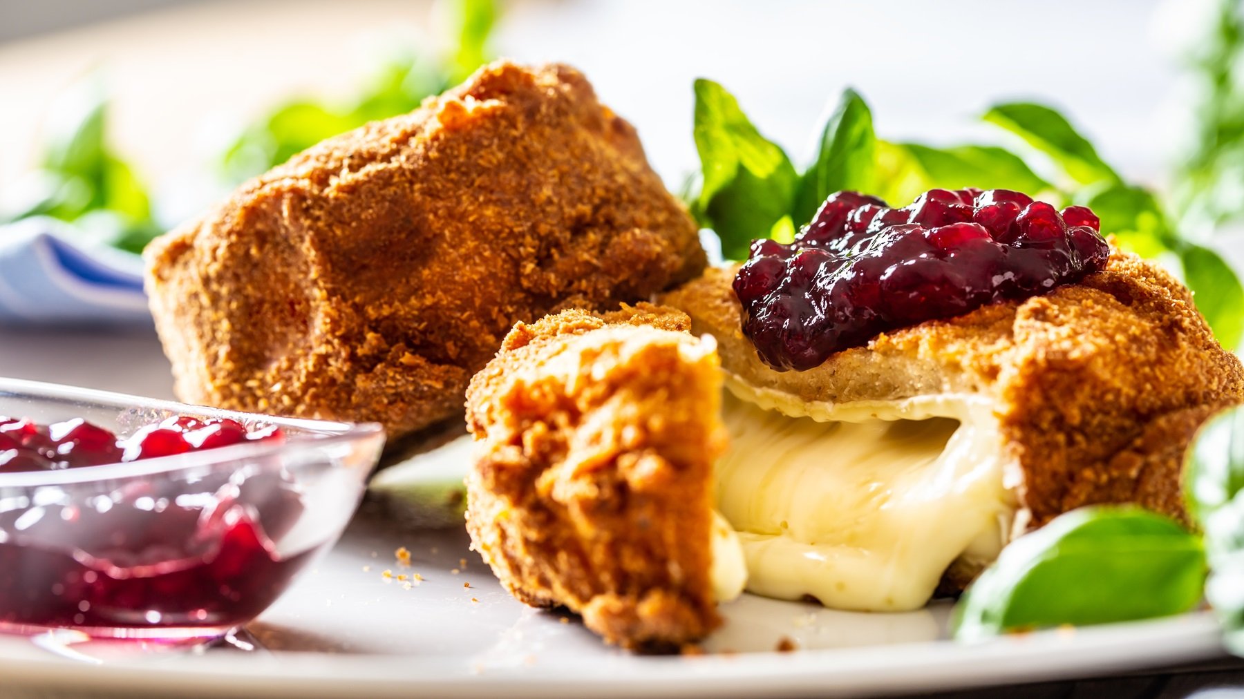 Nahaufnahme vom angeschnittenen Back-Camembert, mit Preiselbeeren garniert. Alles von der Seite und vor hellem Hintergrund fotografiert. Links daneben noch ein kleines Schälchen mit Preiselbeeren und unscharf ein paar grüne Kräuter im Hintergrund.