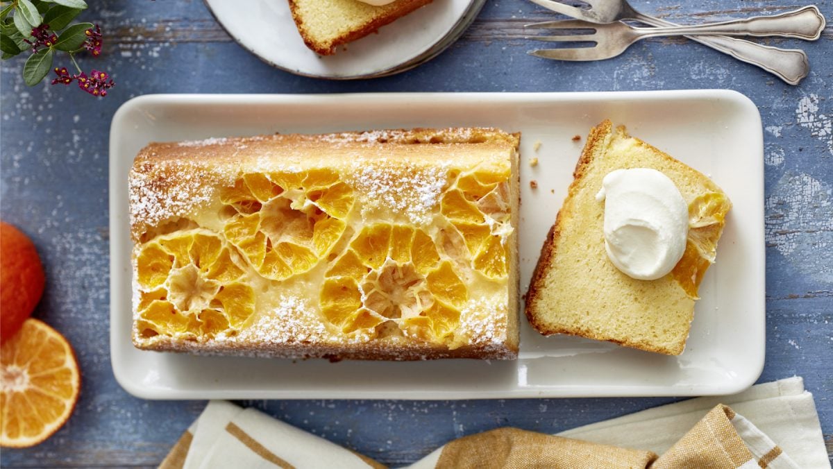 Draufsicht: Angeschnittener gestürzter Clementinenkuchen als Kastenkuchen auf eckigem Teller mit Sahne. Drum herum Besteck und Deko auf blauem Untergrund.