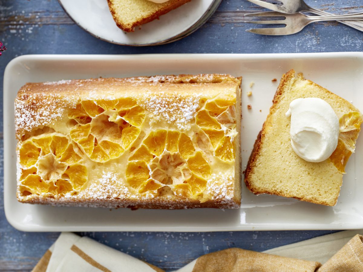 Draufsicht: Angeschnittener gestürzter Clementinenkuchen als Kastenkuchen auf eckigem Teller mit Sahne. Drum herum Besteck und Deko auf blauem Untergrund.