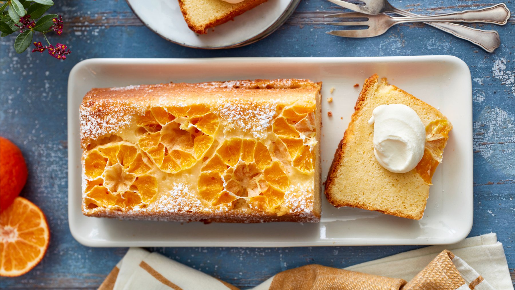 Draufsicht: Angeschnittener gestürzter Clementinenkuchen als Kastenkuchen auf eckigem Teller mit Sahne. Drum herum Besteck und Deko auf blauem Untergrund.