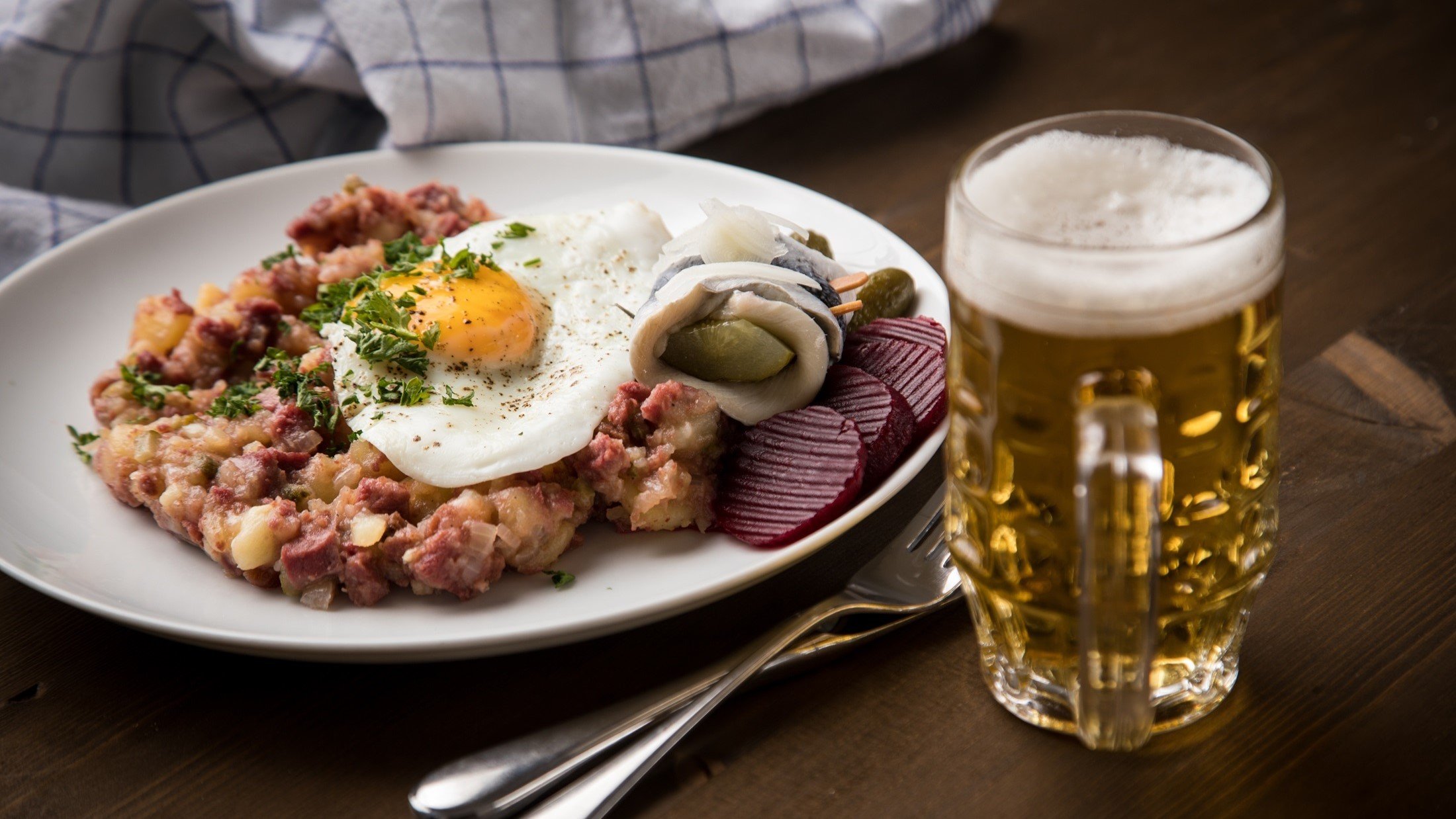 Draufsicht, schräg: Ein Teller mit Hamburger Labskaus steht auf einem Holztisch. Daneben liegt ein Besteck. Neben den steht ein Glas Bier.