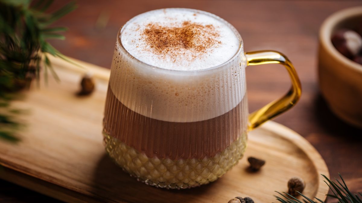 Ein Glas mit dem Eierlikör-Macchiato auf einem hellen Holzbrett von schräg oben fotografiert. Im Hintergrund winterliche Dekoration.