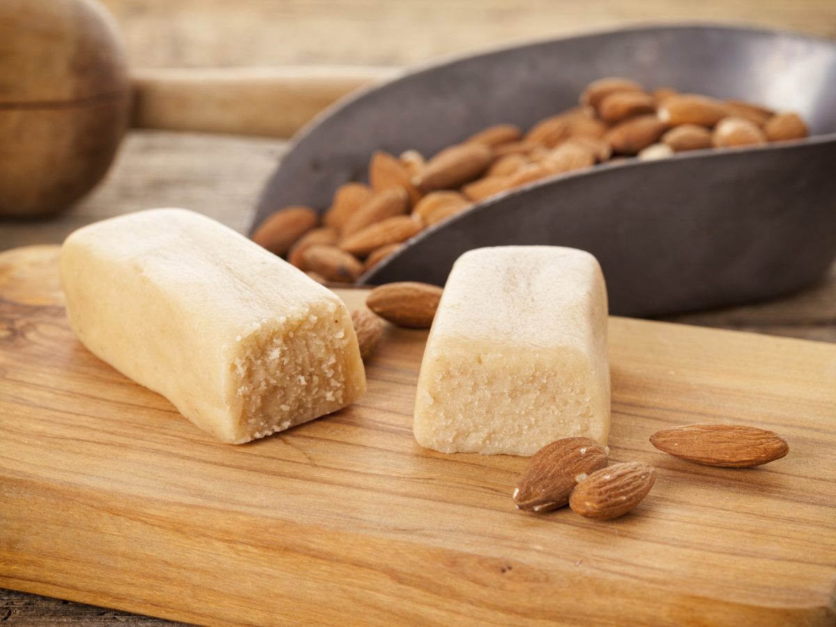 Heller Holztisch mit Marzipan und frischen Mandeln auf einem Holzschneidebrett. Alles nah und von der Seite fotografiert.