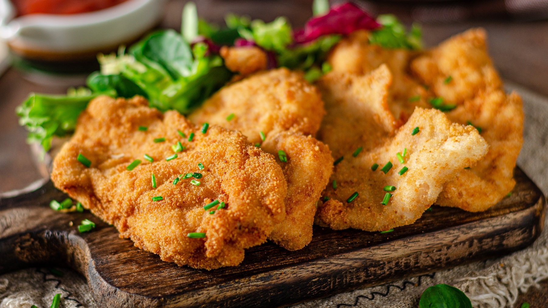 Das Münchner Schnitzel auf einem dunklen Holzbrett und dunklem Holztisch. Alles von der Seite fotografiert mit Salat und Co im Hintergrund.