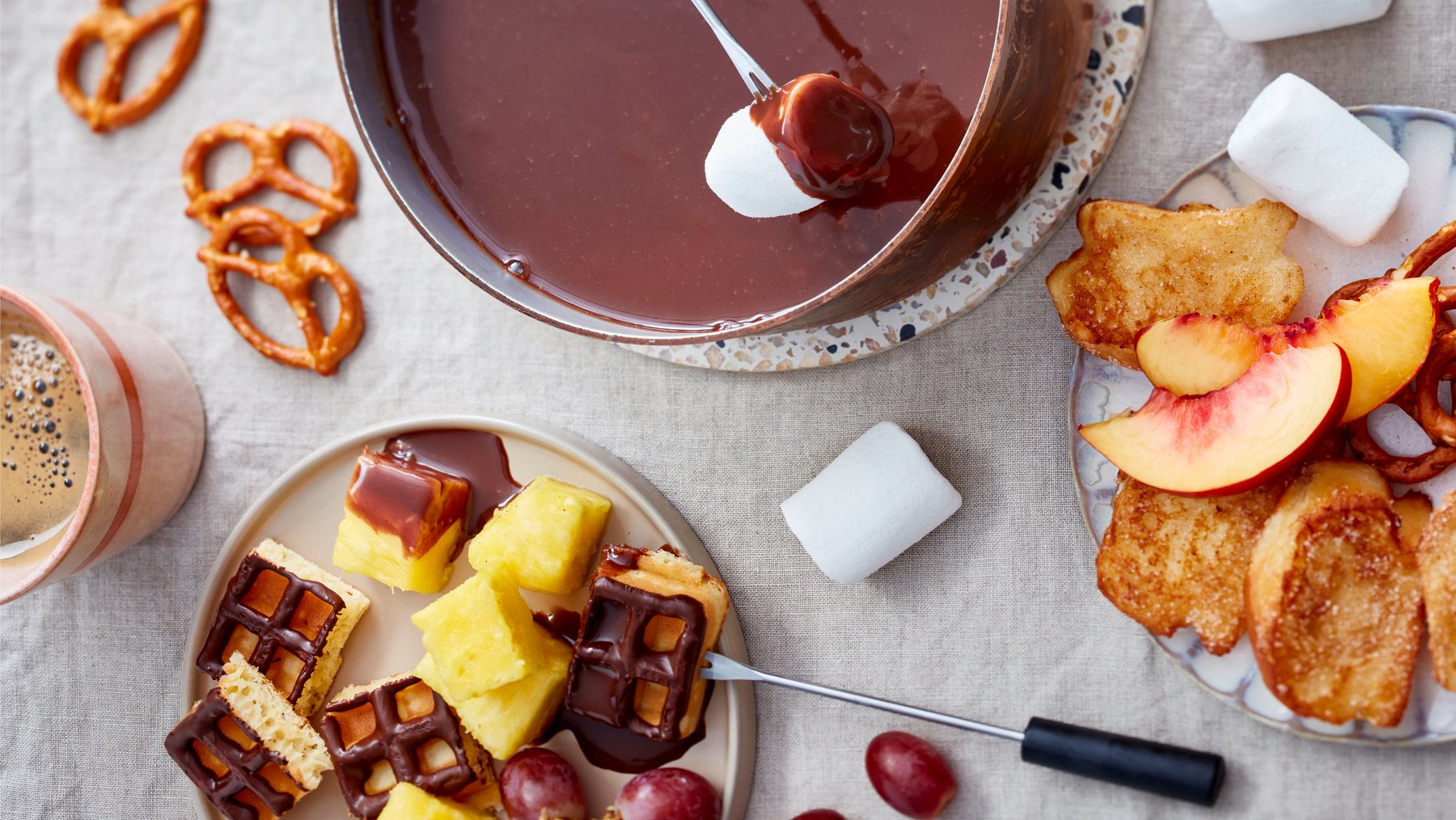 Draufsicht: In zwei Schalen Nougatfondue mit armen Ritterlein, drum herum Knusperbrezeln, Obst, Marshmallows und Mini-Waffeln