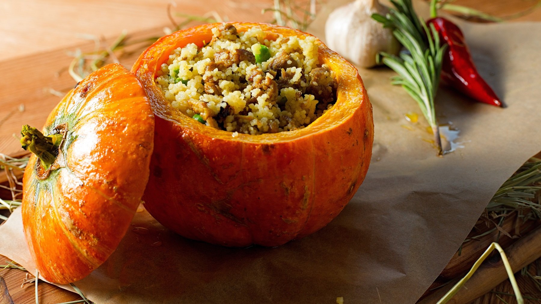 Ein Holztisch mit dem Kürbis auf Backpapier rund vielen Kräutern wie Rosmarin, Chili und Knoblauch. Alles von der Seite fotografiert.