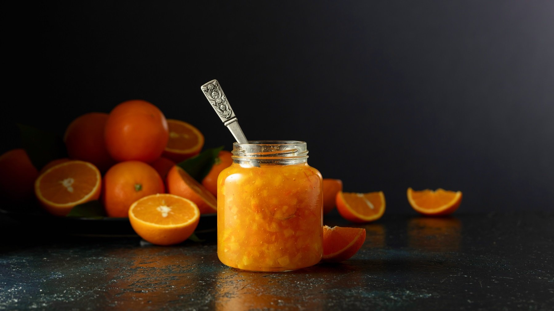 Ein Glas mit der Orangenmarmelade und ein paar halbierten Orangen auf dunklem Untergrund und vor schwarzem Hintergrund.