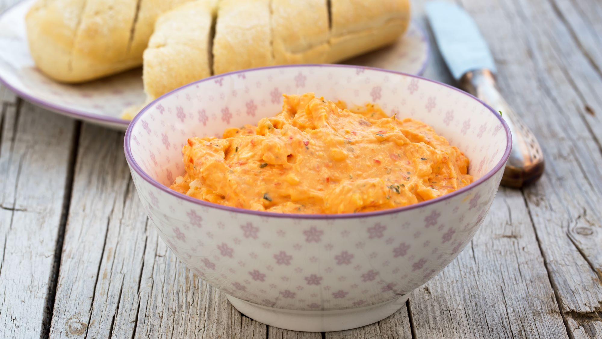 Selbstgemachter Paprika-Feta-Dip mit Kräutern in dekorativer Schale, serviert auf einer Holzoberfläche mit Brotscheiben.