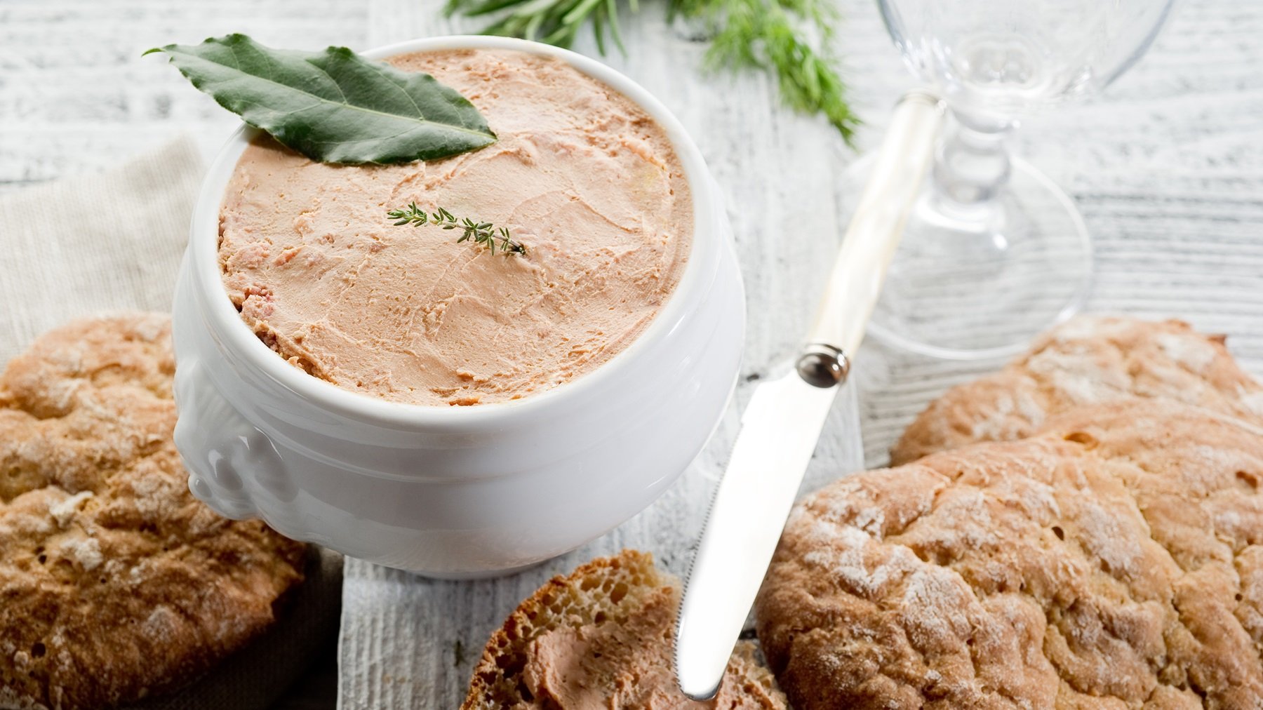 Draufsicht auf ein weißes Porzellangefäß mit Pâté di fegato, darauf ein Lorbeerblatt. Daneben frischen Brot und ein Messer.