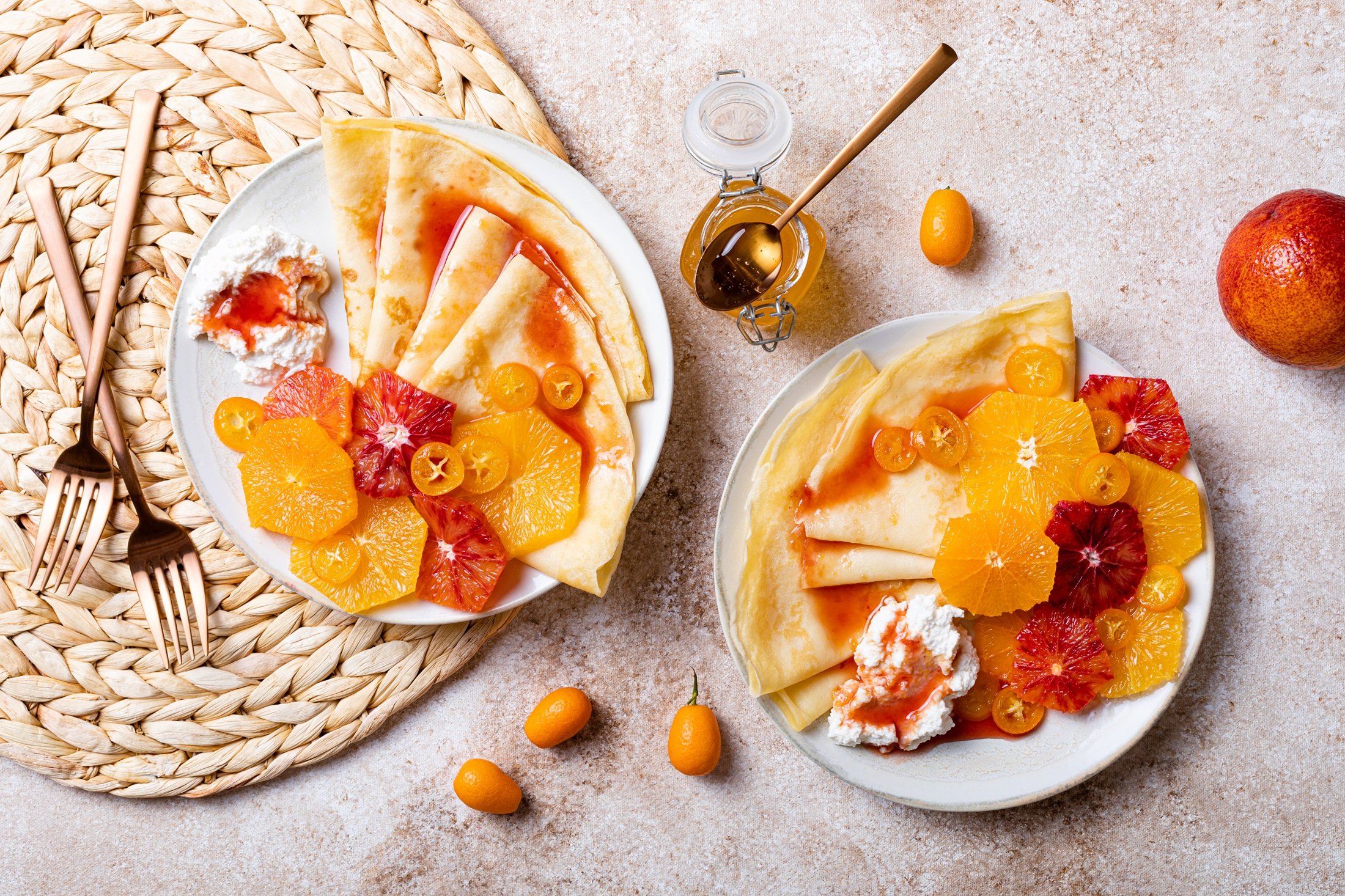 Draufsicht: Zwei weiße Teller mit Pfannkuchen und karamellisierten Orangen und einem Klecks Sahne. Drum herum liegen kleine Kumquats sowie zwei vergoldete Gabeln und ein vergoldeter Löffel.