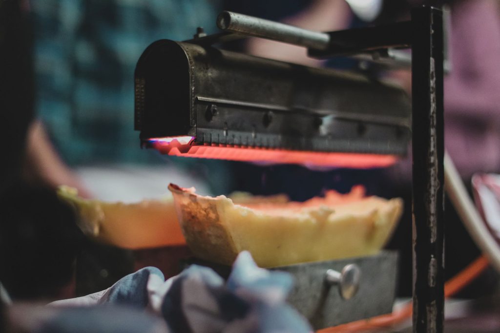 Ein halber Raclettekäse schmilzt unter einem glühenden Grill im sanften Licht vor unscharfem Hintergrund.