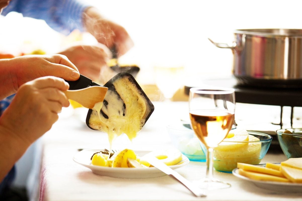 Eine Hand schabt geschmolzenen Käse aus einer Raclette-Pfanne auf einen Teller mit Kartoffeln, daneben ein Glas Weißwein.