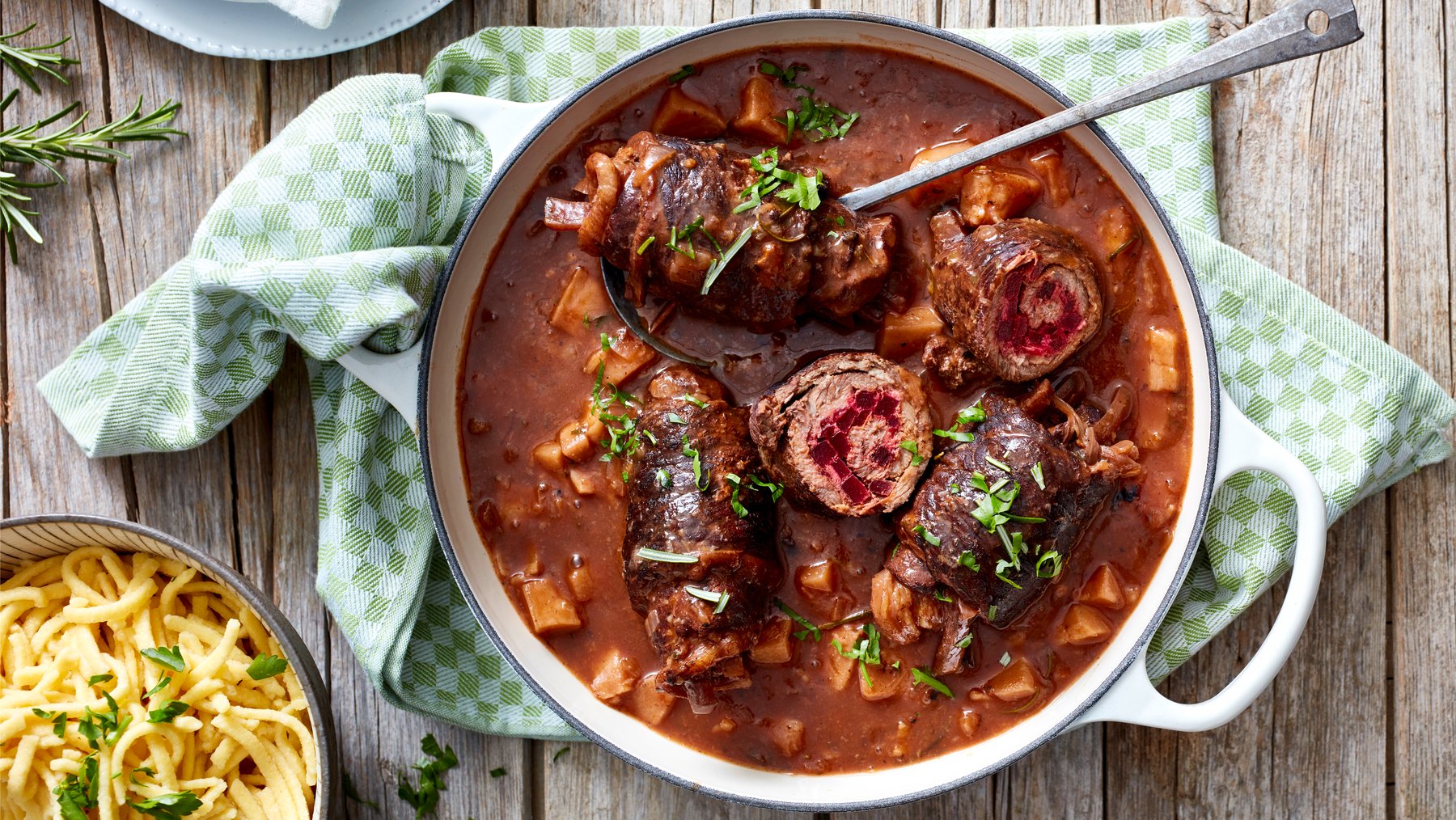 Draufsicht: Rinderroulade mit Rote-Bete-Füllung in einem Topf mit brauner Sauce, daneben Spätzle mit hellem, rustikalen Hintergrund.