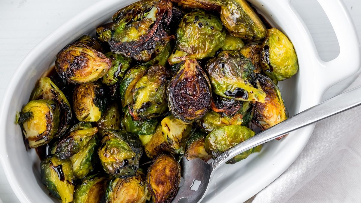 Eine weiße Auflaufform mit dem Rosenkohl aus dem Airfryer und einem silbernen Löffel auf einem weißen Tisch. Alles sehr nah und direkt von oben fotografiert.