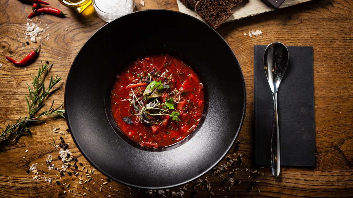 Ein schwarzer Teller mit der Suppe auf einem braunen Holztisch direkt von oben fotografiert. Daneben noch Löffel, Gewürze und Serviette in rustikalem Ambiente.