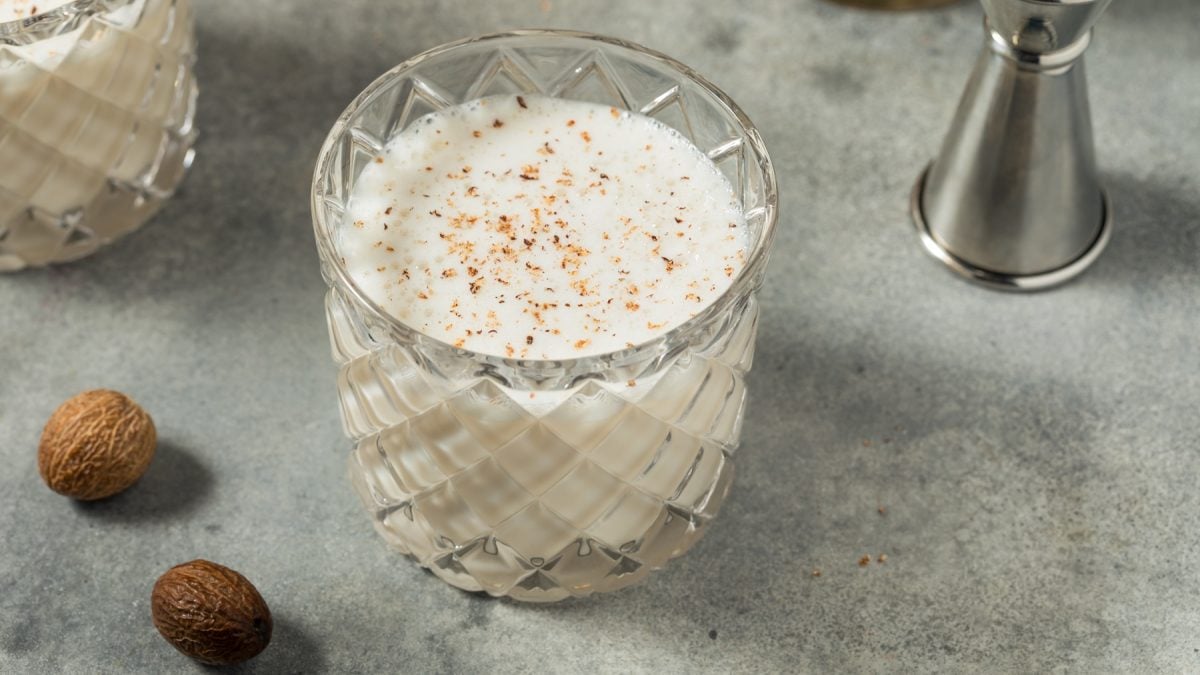 Glas mit Rum Milk Punch vor grauem Hintergrund, daneben frische Muskatnüsse.