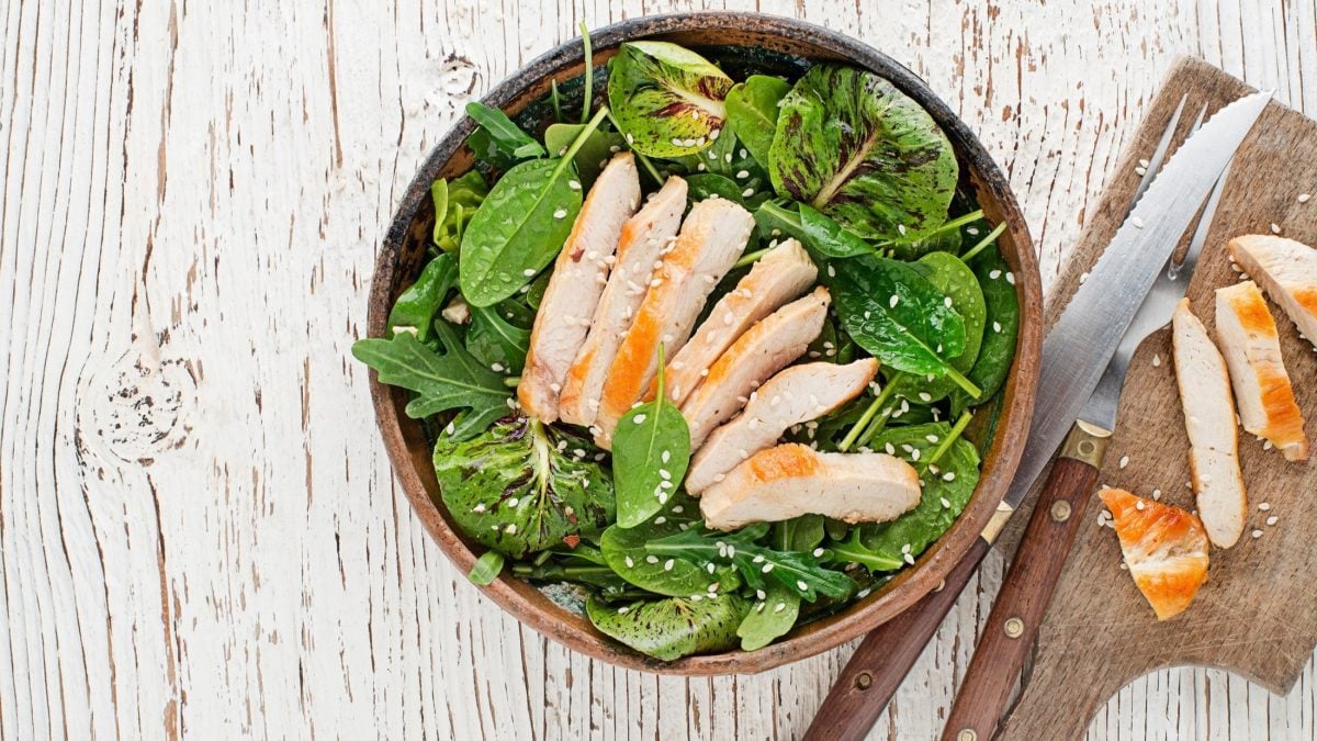 Eine Schüssel mit einem Salat mit Hähnchenbrust und Mandel-Dressing steht auf einem hellem Untergrund aus Holz. Daneben liegt ein Holzbrettchen mit Besteck. / Draufblick