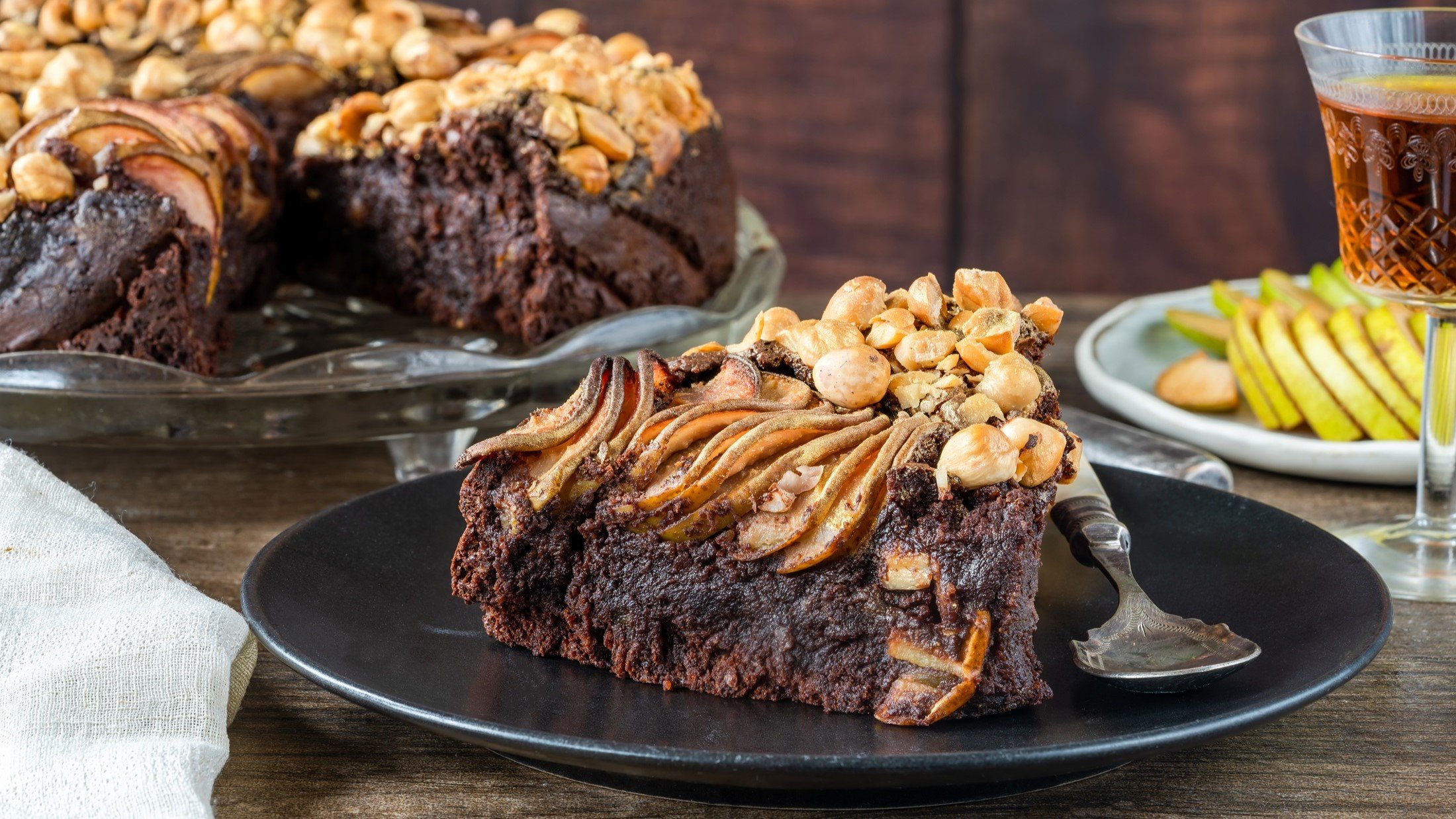 Frontal: eine angeschnittene Schokoladen-Torte mit Nüssen und Äpfeln. Sowie ein Teller mit einem Stück Torte. Im Hintergrund sind geschnittene Äpfel und ein Glas Tee.