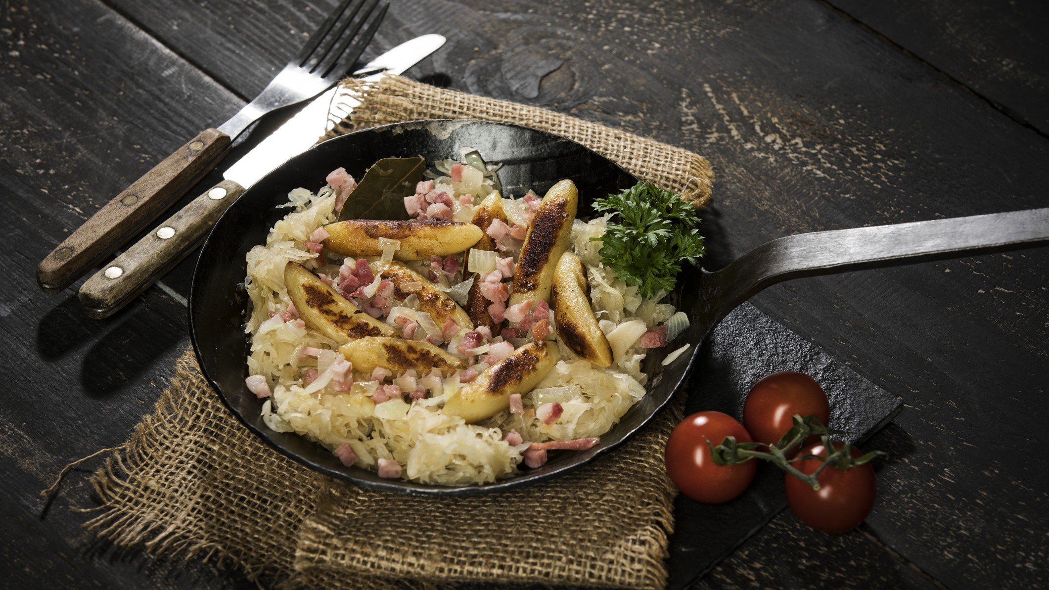 Schupfnudeln mit Sauerkraut in einer rustikalen Pfanne, die auf einem rustikalen Tisch aus Holz steht. Neben der Pfanne liegen Besteck sowie frische Tomaten.