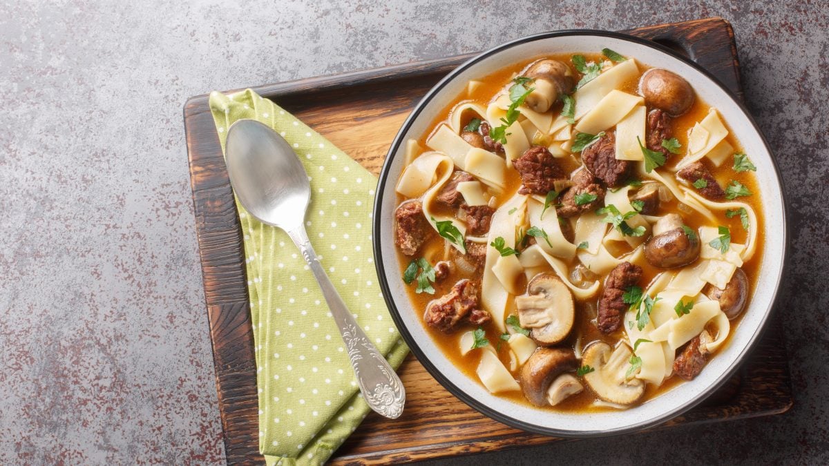 Weißer Teller gefüllt mit einer Stroganoff-Nudelsuppe. Dieser steht auf einem Holzbrett, auf dem auch ein Löffel und eine grüne Serviette liegt.