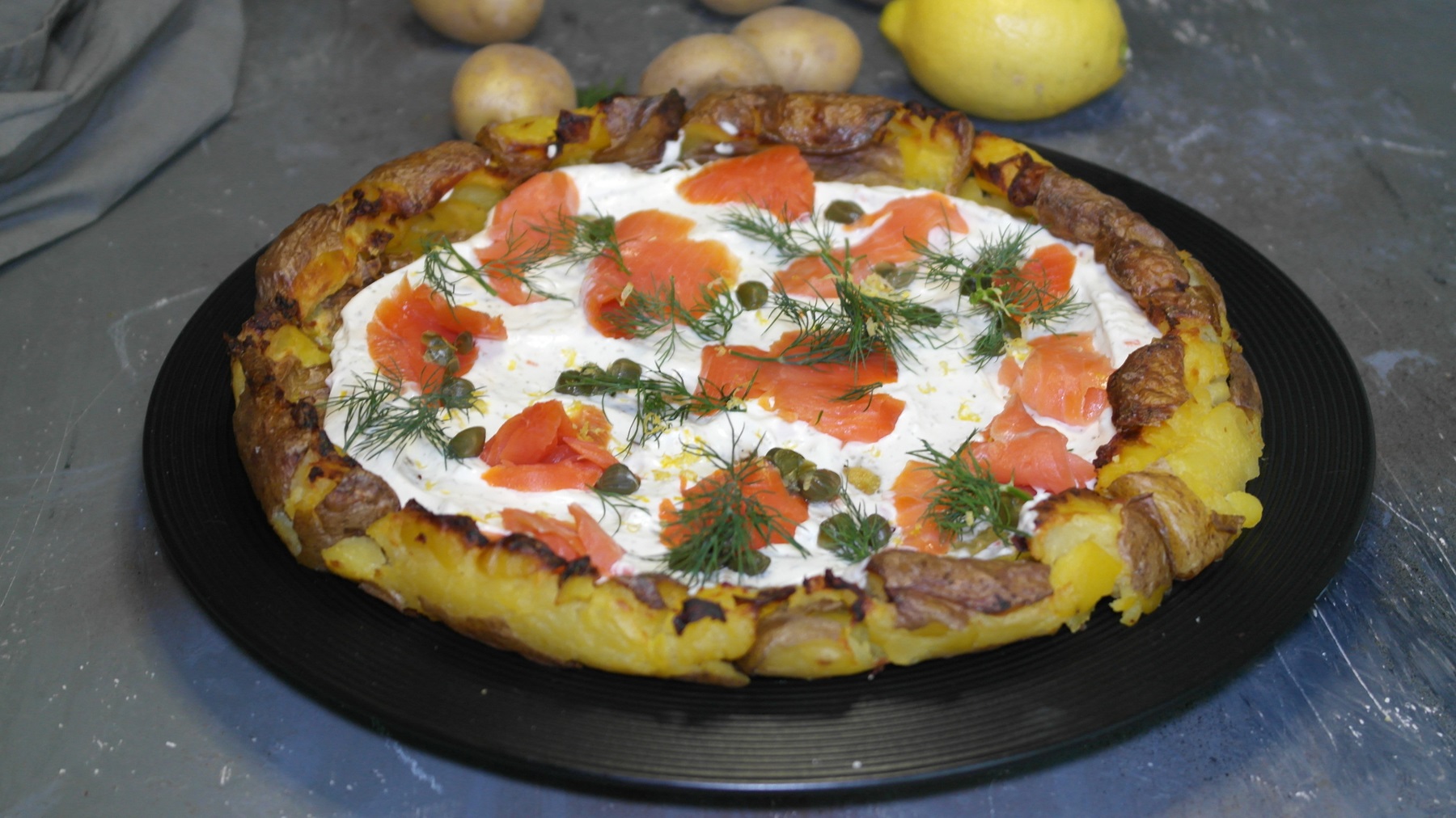 Ein schwarzer Teller, mit der Kartoffelpizza belegt mit Lachs, Kapern, Dill und Zitronenabrieb. Alles von leicht schräg oben auf grauem Steinuntergrund fotografiert.