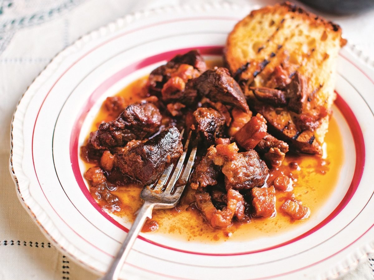 Ein weißer Teller mit roter Verzierung und darin das köstliche Gulasch mit einer gerösteten Weißbrotscheibe. Alles von schräg oben auf einem weißen Tisch fotografiert.