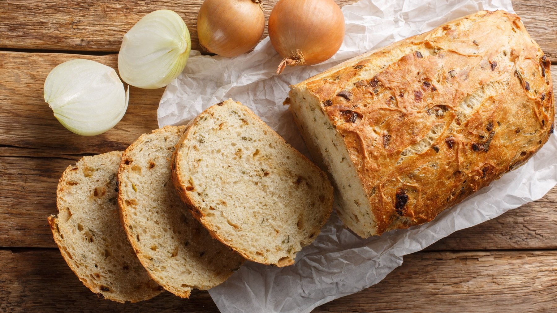 Draufsicht auf ein angeschnittenes Zwiebelbrot, daneben eine halbierte und eine ganze frische Zwiebel.