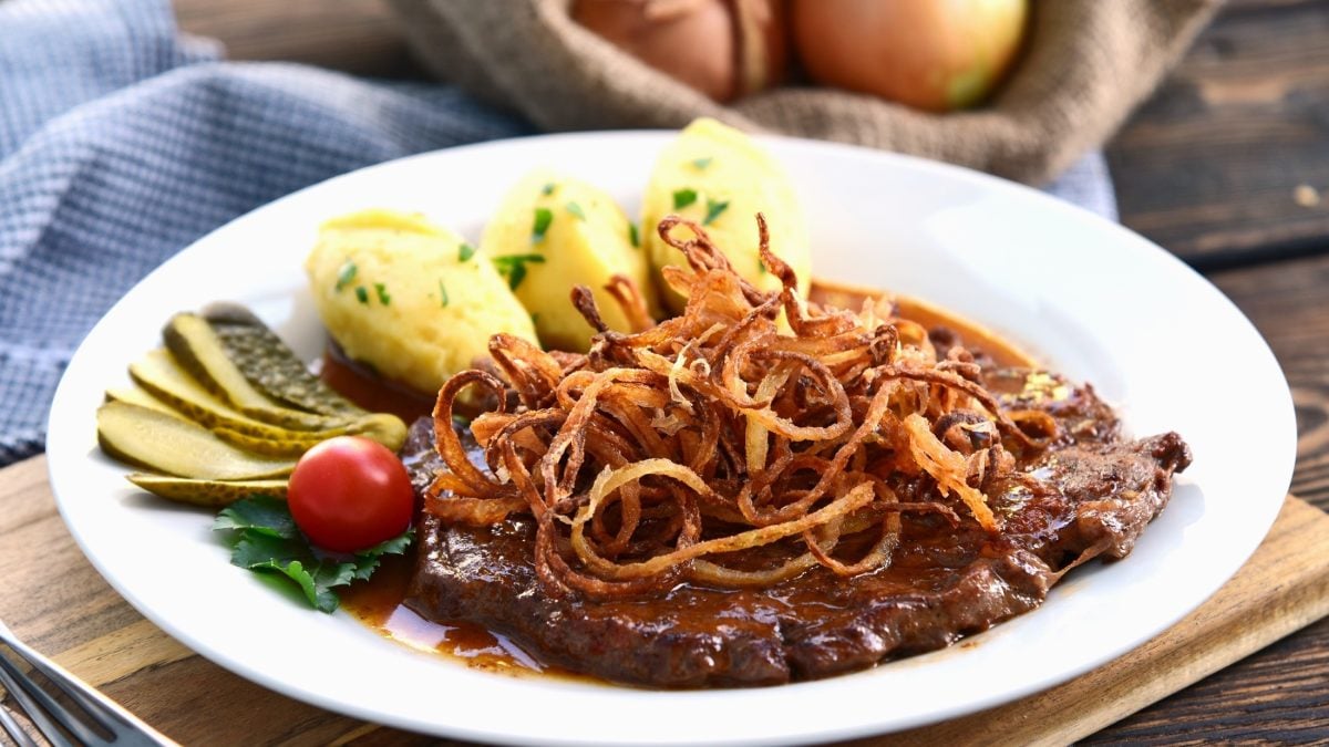 Ein Zwiebelrostbraten mit Sauce und Beilagen auf einem hellem Teller aus Porzellan. / Draufsicht, schräg.