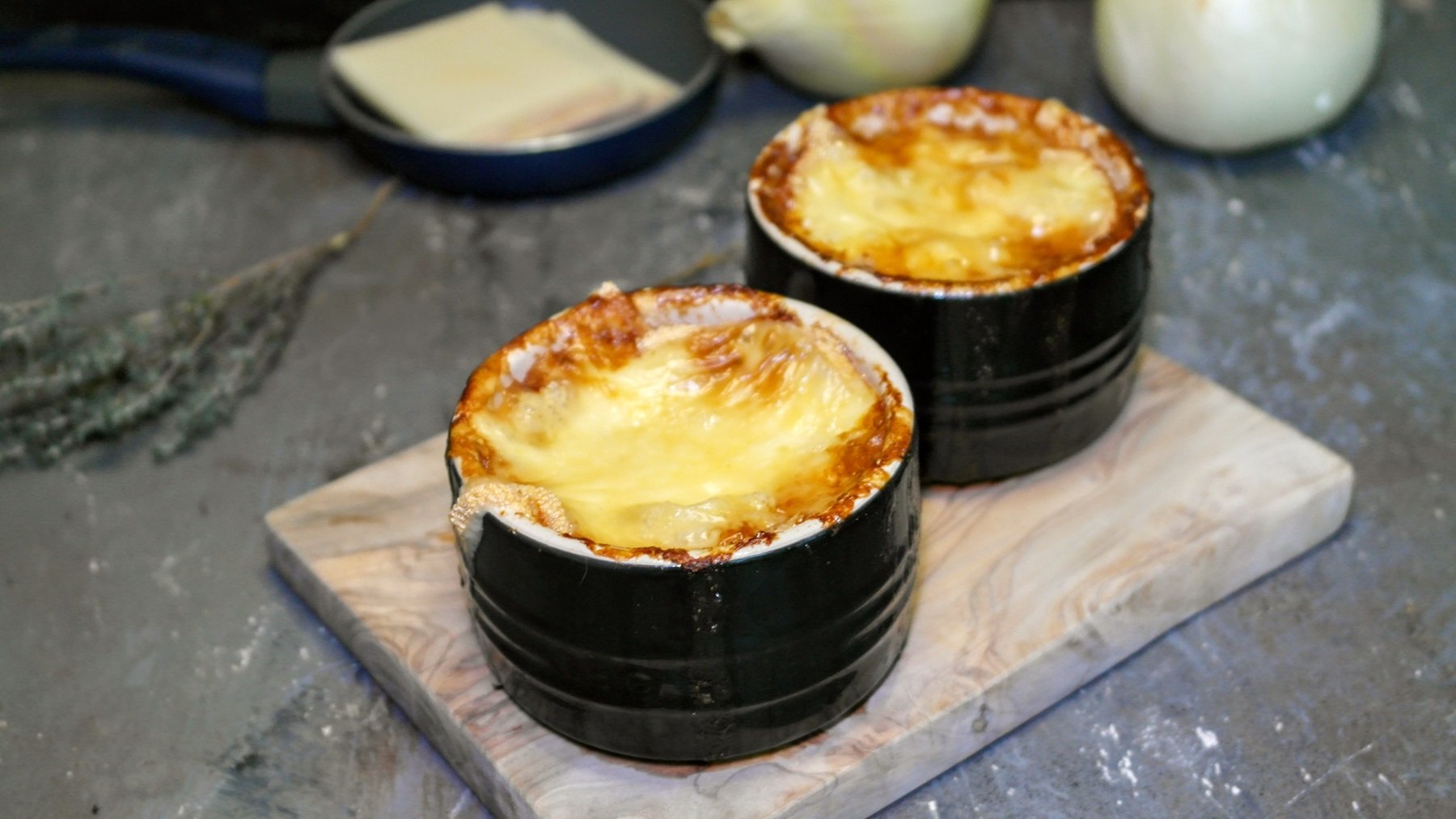 Zwei schwarze Schälchen mit der überbackenen Zwiebelsuppe auf einem kleinen Holzbrett und Steinuntergrund. Alles von schräg oben fotografiert.