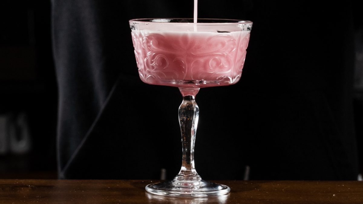 Pink Gin Sour wird in ein elegantes, verziertes Cocktailglas aus Glas mit filigranem Stiel auf dunklem Hintergrund gegossen.