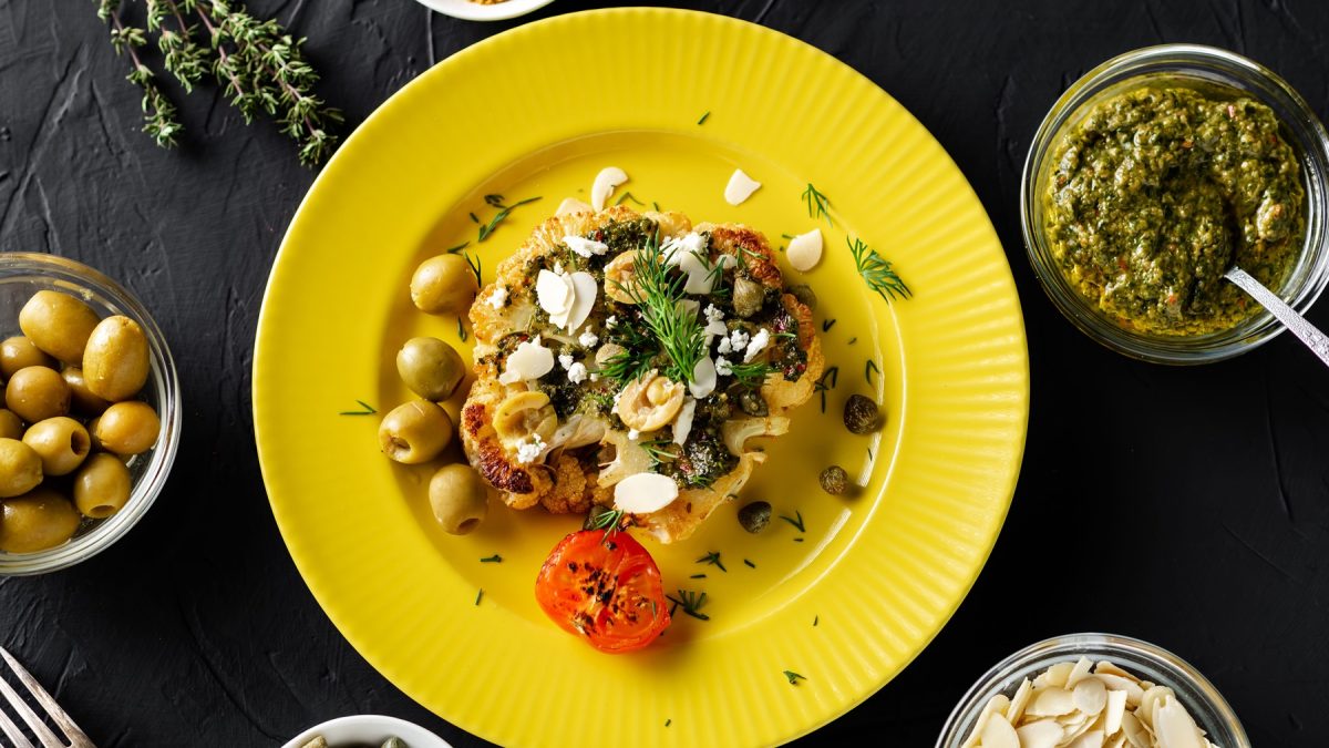 Elegantes Gericht mit Blumenkohlsteak, grünen Oliven und frischen Kräutern auf gelbem Teller, begleitet von Schälchen mit Pesto und Mandeln.