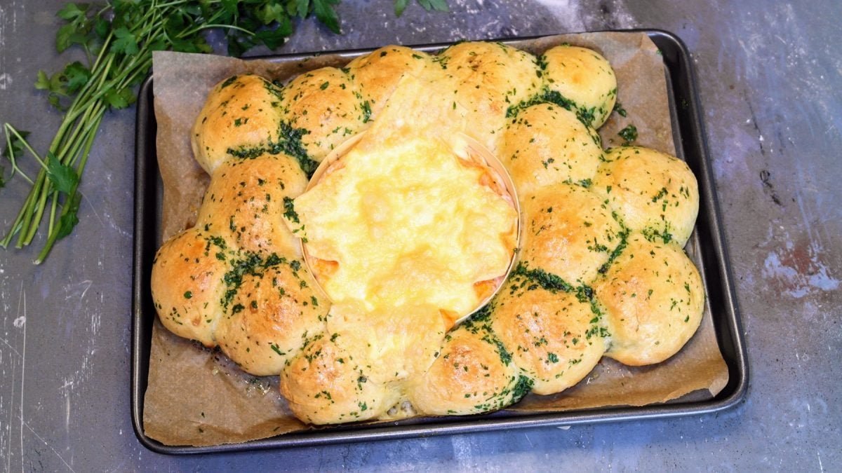 Ein Blech mit dem Brötchenkranz und Ofenkäse auf einem steinernen Tisch von schräg oben fotografiert. Im Hintergrund unscharf ein Bund Petersilie erkennbar.