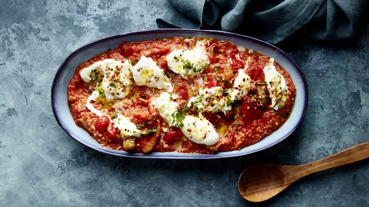 Ovaler Teller mit Büffelmozzarella auf Röstgemüse à la Romesco, daneben ein Holzlöffel auf blaugrauem Untergrund.