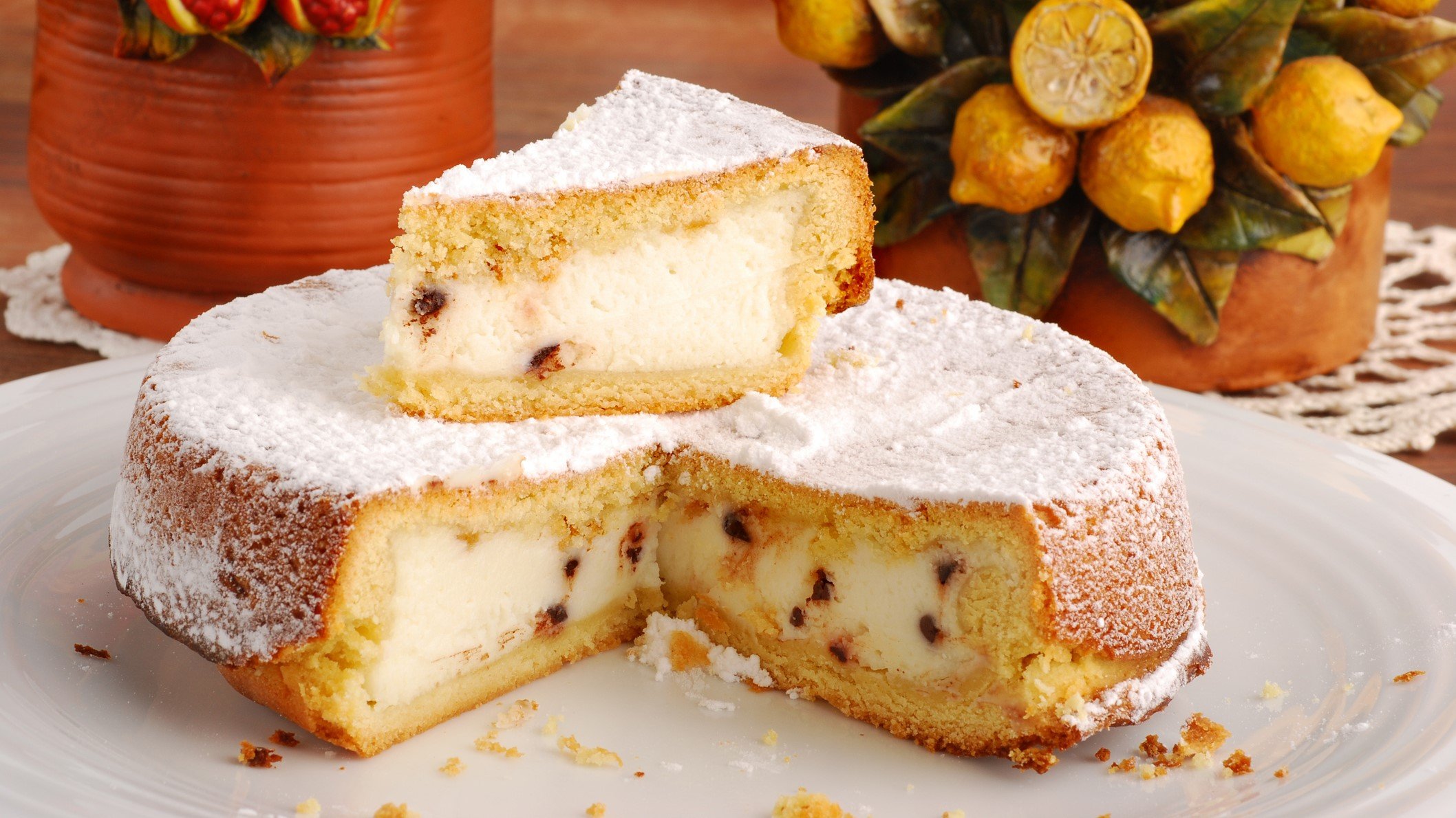 Ein angeschnittene Cassata al forno mit Schokoladenstückchen und Puderzucker steht auf einem weißen Teller, im Hintergrund dekorative Keramiktöpfe.