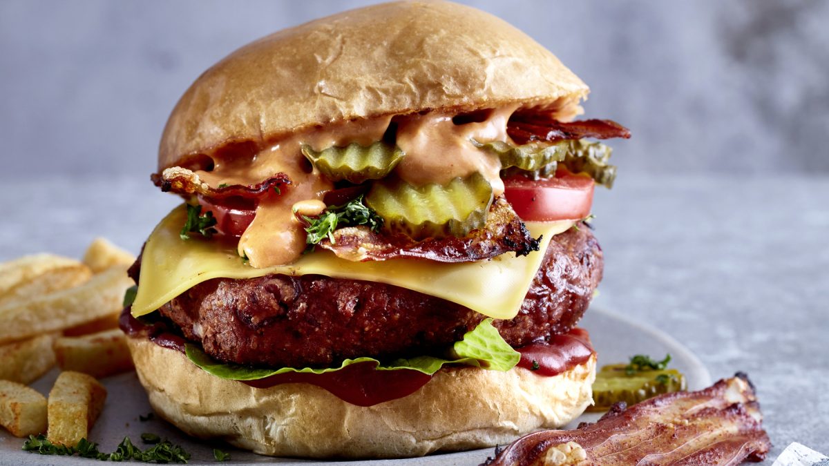 Cheesy Böhnchenburger mit Pommes vor grauem Hintergrund, dazu Pommes und veganer Bacon.