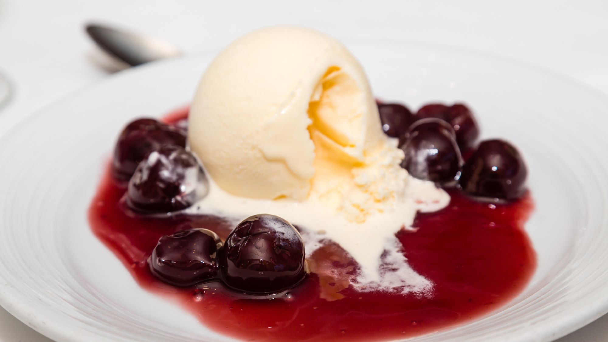 Cherries Jubilee mit Vanilleeiskugel und eingelegten Kirschen in süßer roter Sauce, serviert auf einem eleganten weißen Teller.