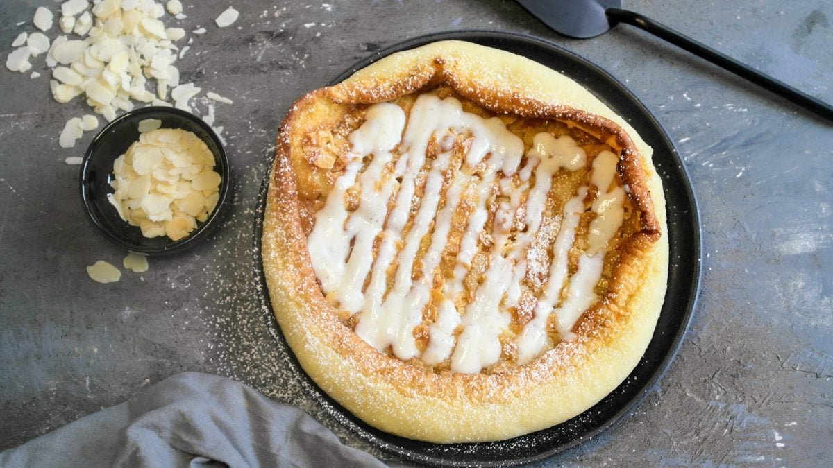 Ein schwarzer Teller mit dem Bienenstich-Pfannkuchen auf steinernem Untergrund, mit einem grauen Tuch und einem Schälchen mit Mandelblättchen.