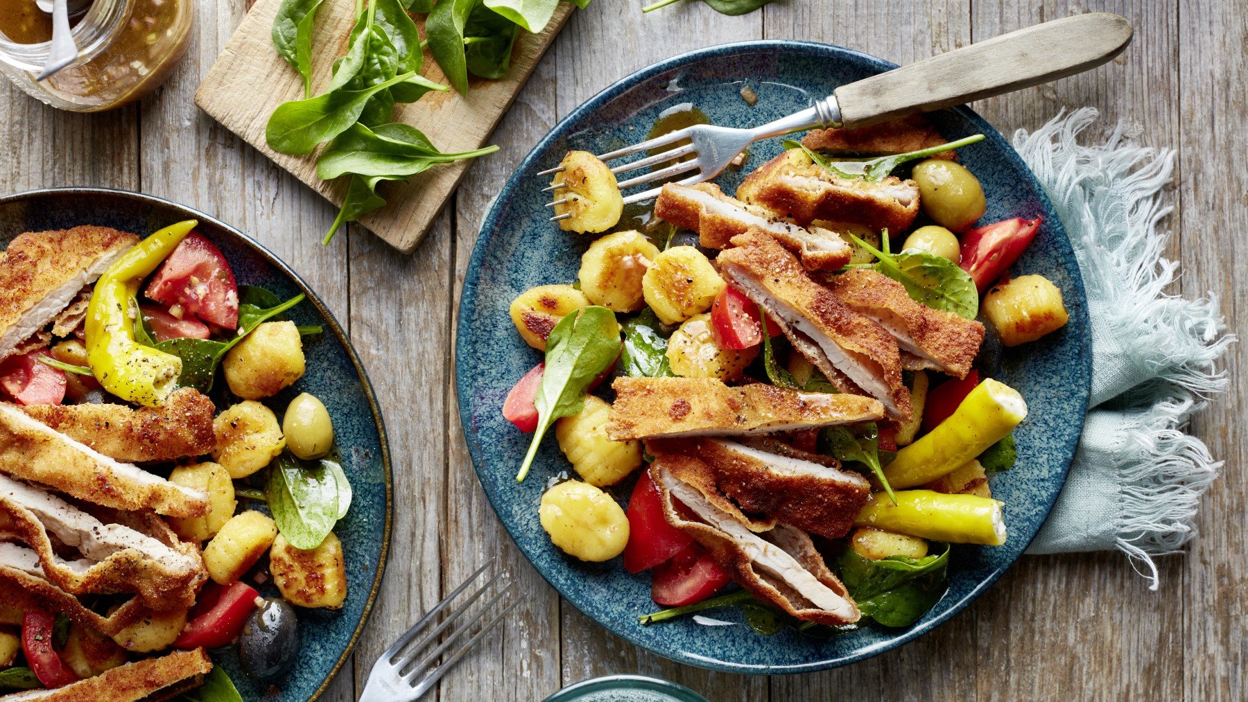 Antipasti-Schnitzel-Salat auf zwei dunkelblauen Tellern verteilt auf hölzernem Hintergrund.