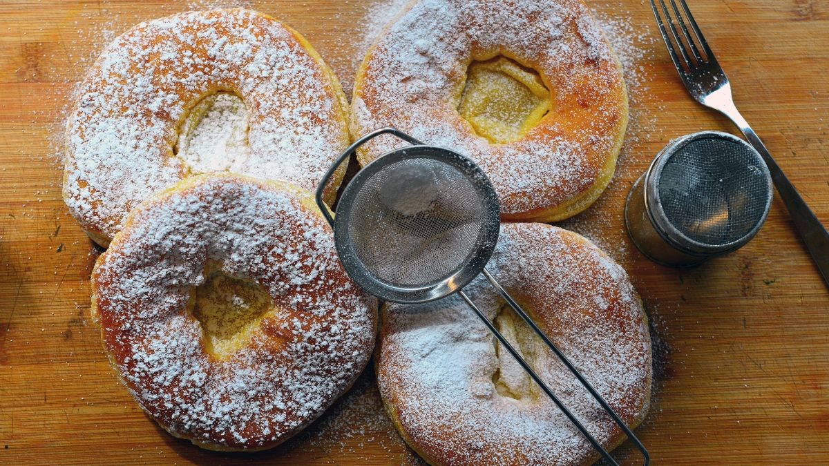 Draufsicht: Auf einem Holzbrett liegen vier Ausgezogene. Das Schmalzgebäck ist mit Puderzucker bestreut.