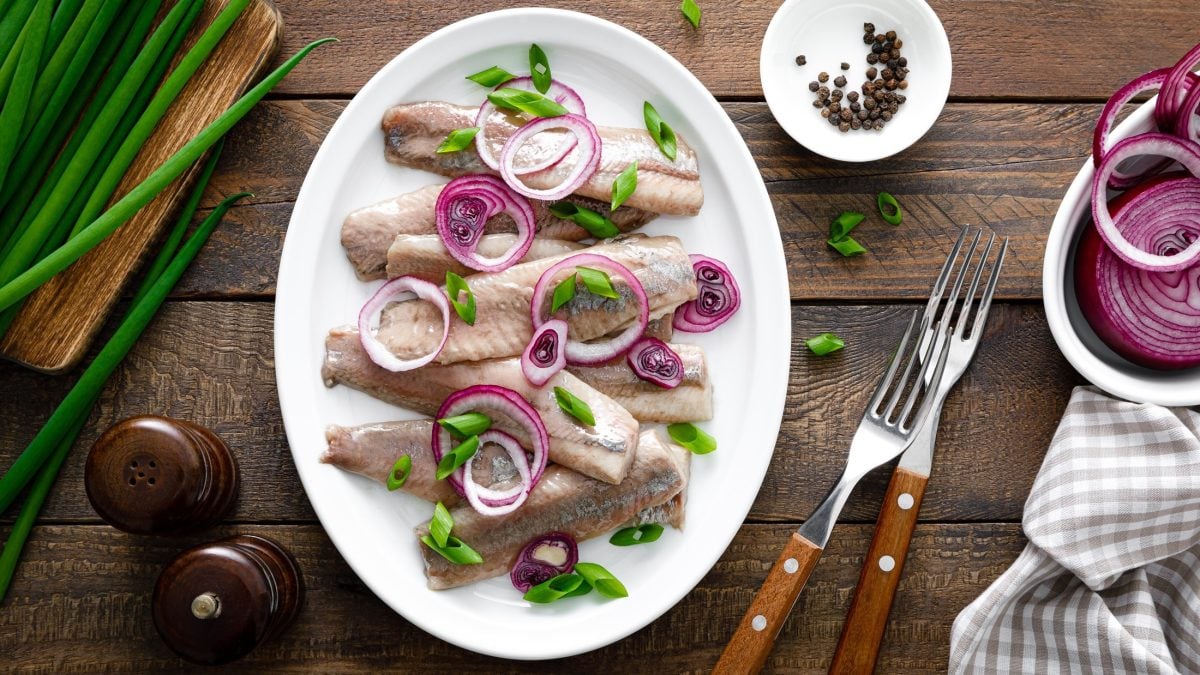 Draufsicht: Mehrere Fischfilets, beziehungsweise Bismarckheringe, auf einem hellen Teller. Der steht auf einem rustikalen Holztisch. Drum herum liegen Bestecke, Frühlingszwiebeln, Gewürzmühlen und ein Tuch.