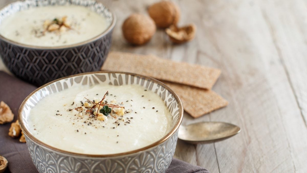 Draufsicht, schräg: Zwei Schüsseln mit einer Blumenkohlsuppe mit Walnuss auf einem rustikalen Untergrund aus Holz. Drum herum liegen ein silberner Löffel und Walnüsse.