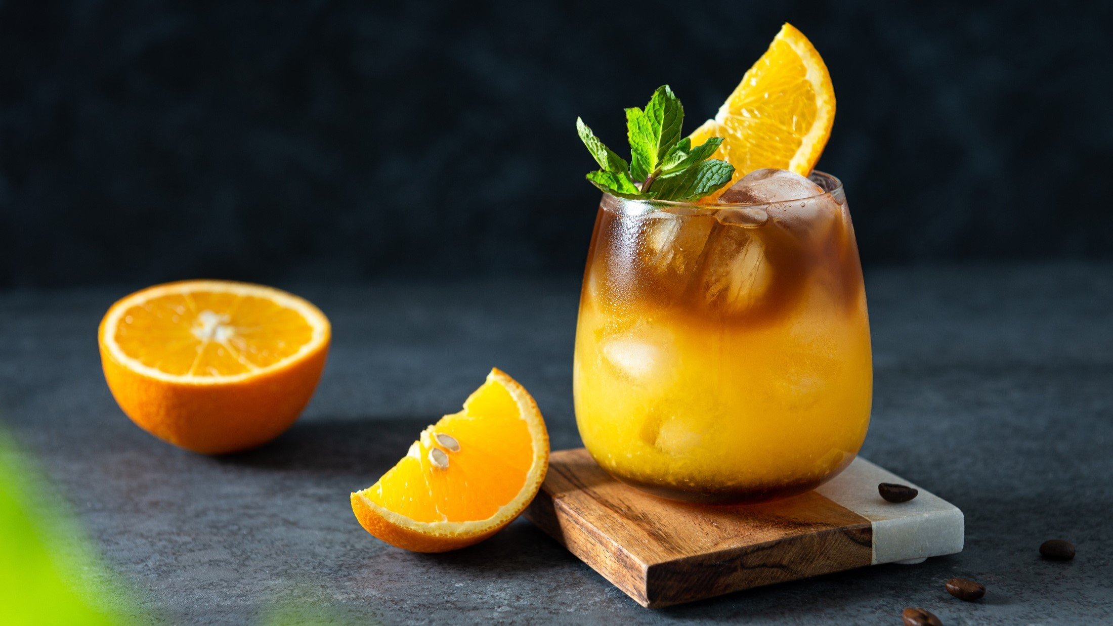 Frontal: Ein Glas mit einem Getränk aus Kaffee und Orangensaft in einem Glas. Es ist mit Minzblättern und einer halben Scheibe Orangensaft verzieret. Daneben liegen eine halbe und eine viertel Orange.