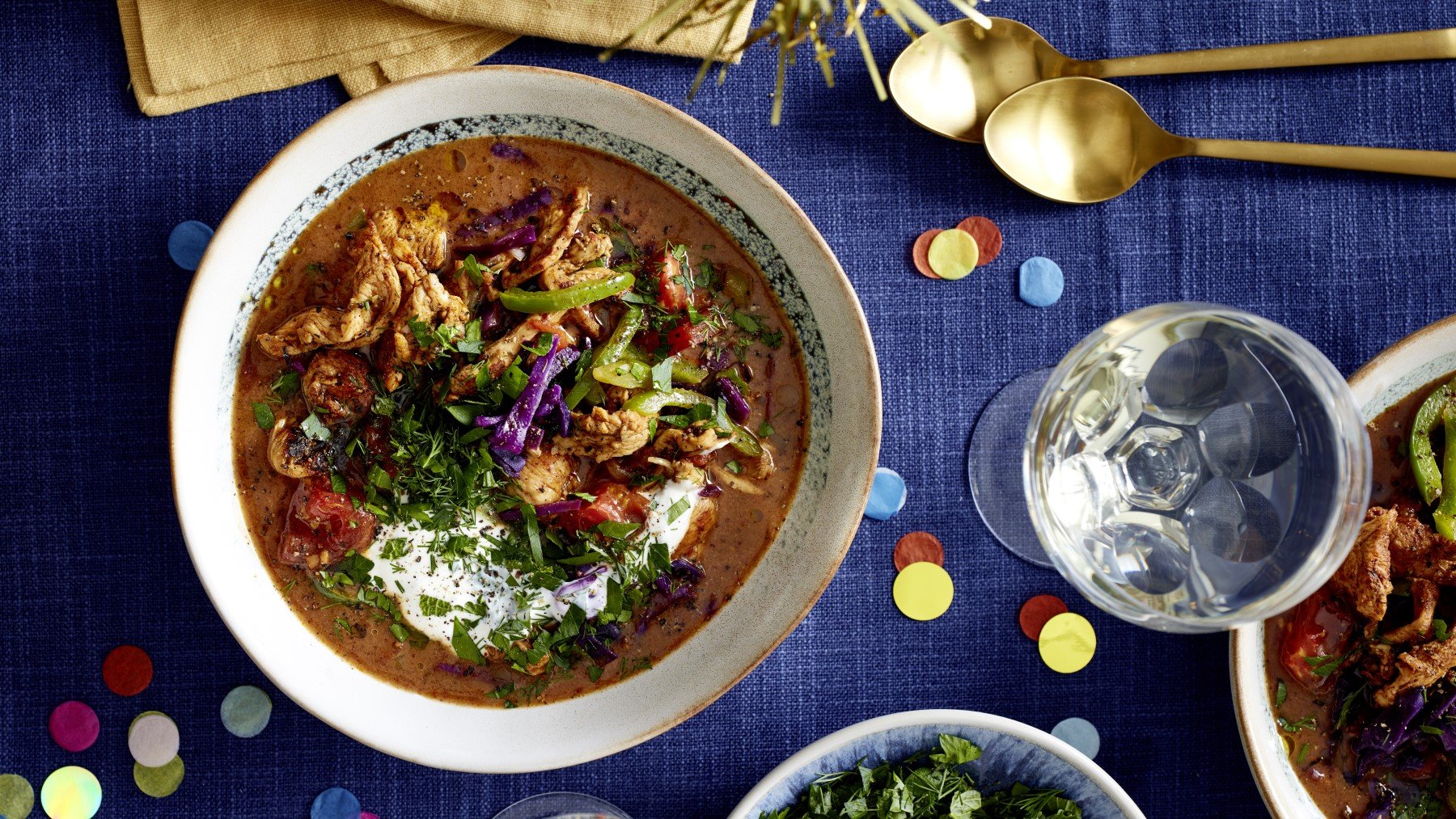 Hähnchensuppe Döner-Art in einer Schüssel auf dunkelblauem Hintergrund. Rechts oben goldene Löffel und Konfetti auf dem Hintergrund verteilt.