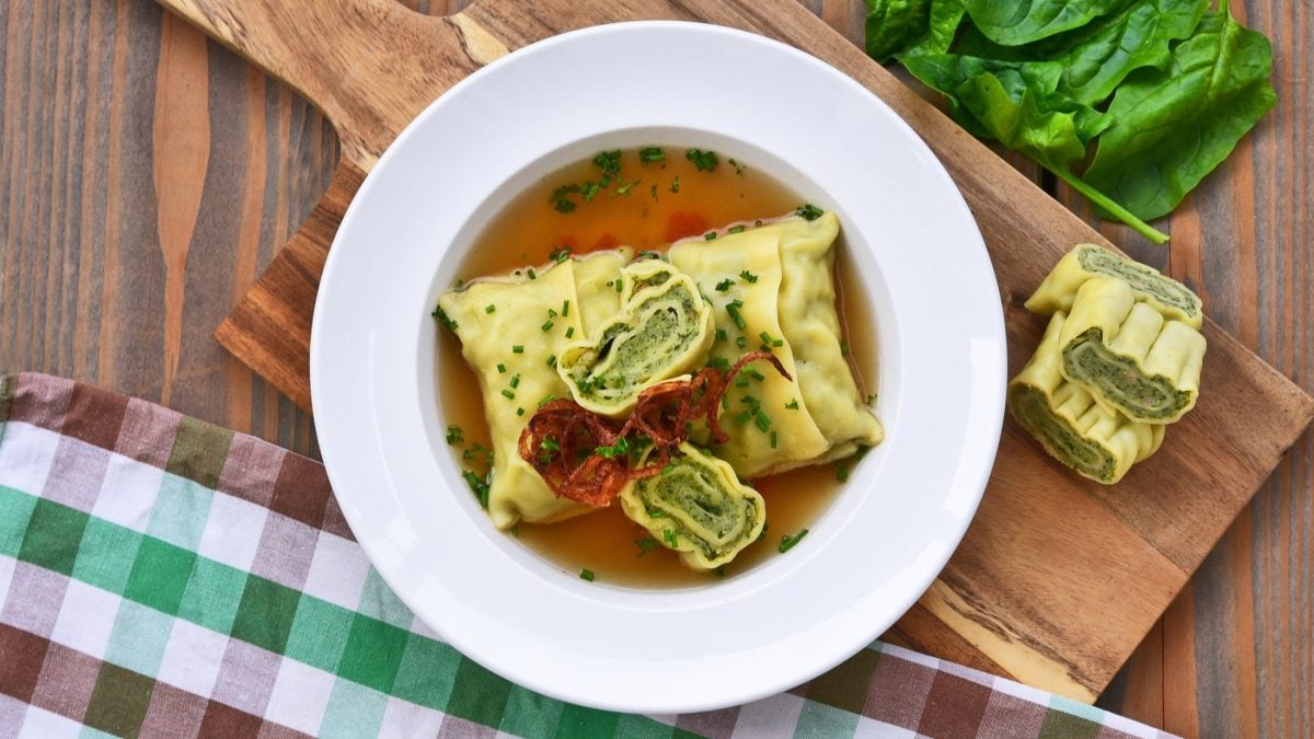 Draufsicht: Ein weißer Teller mit einer Maultaschensuppe mit Spinat auf einem Holzbrett. Drum herum liegen frische Spinatbätter, ein Küchenhandtuch und zwei angeschnittene Maultaschen.