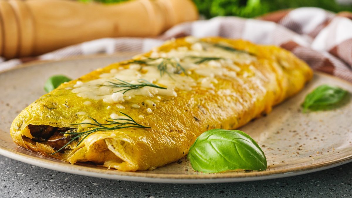 Frontal Ein französisches Omelette mit frischen Kräutern, dass zusammen geklappt ist, auf einem Teller. Im Hintergrund liegt eine lange Pfeffermühle aus Holz.