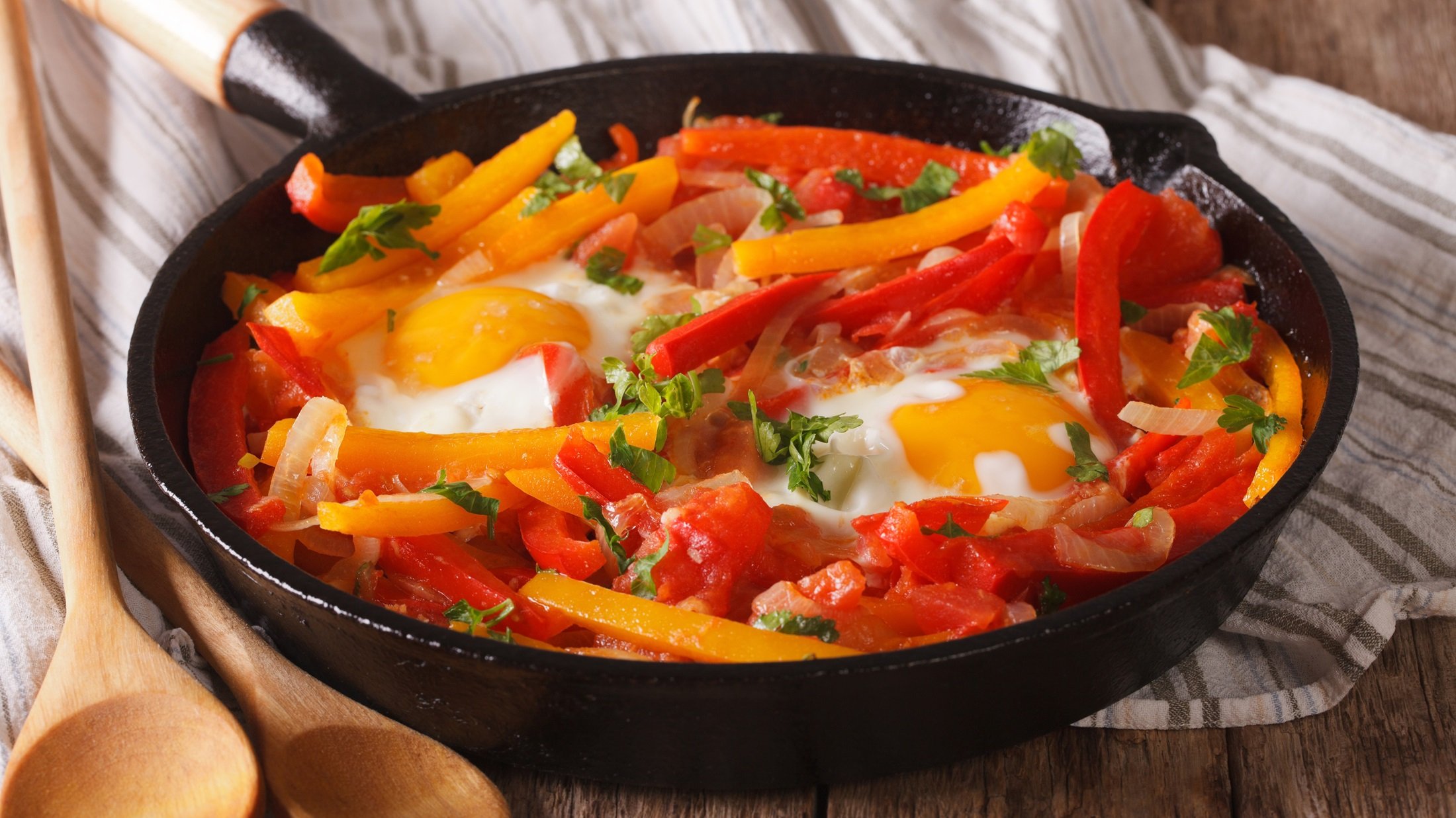 Draufsicht, schräg oben: Eine gusseiserne Pfanne mit Gemüse, wie Paprika, Tomaten und Zwiebeln. In der Mitte sind zwei Spiegeleier.