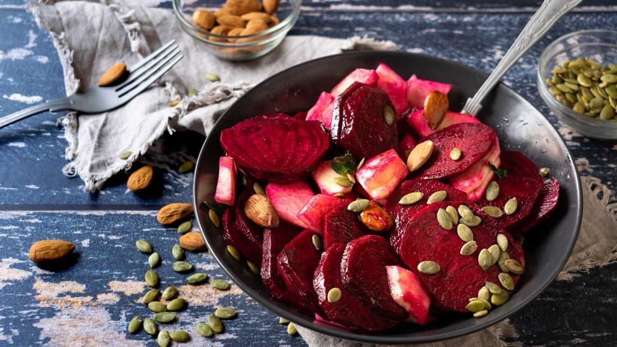 Draufsicht: Ein heller Untergrund auf dem einen Schüssel mit einem Rote-Bete-Salat mit Apfel steht, in dem ein Löffel steckt. Drum herum liegen mandeln mit Haut sowie Kürbiskerne.