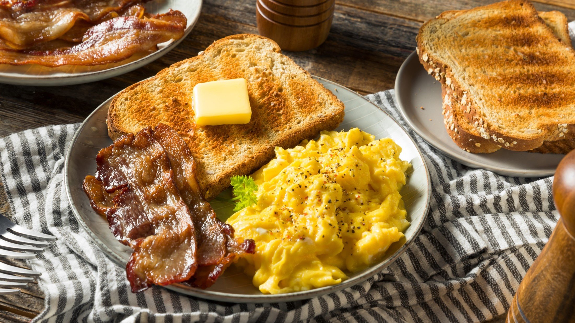 Draufsicht: Ein gedeckter Tisch. Ein großer runder Teller mit Rührei und Speck und einem Toast auf dem Butter liegt. Drumherum ein weiterer Teller mit Brot und noch ein Teller mit gebraten Speckstreifen.