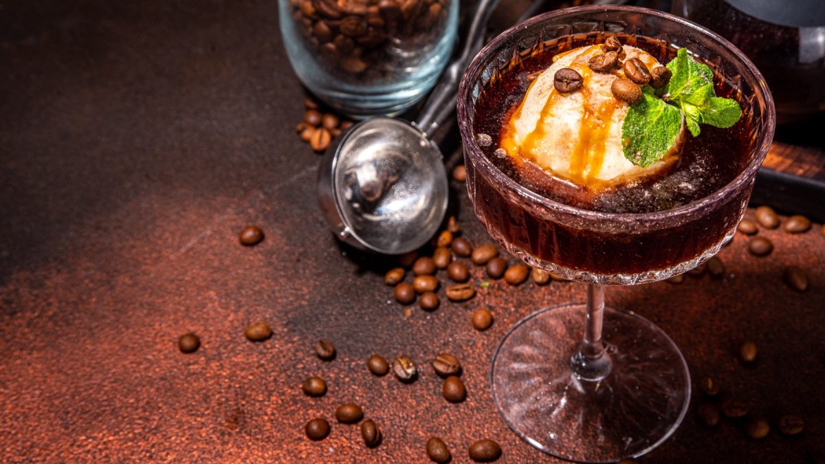 Nahaufnahme eines Glases mit cremigem Espresso Martini Affogato, dekoriert mit Karamellsirup, frischer Minze und Kaffeebohnen, umgeben von verstreuten Bohnen.