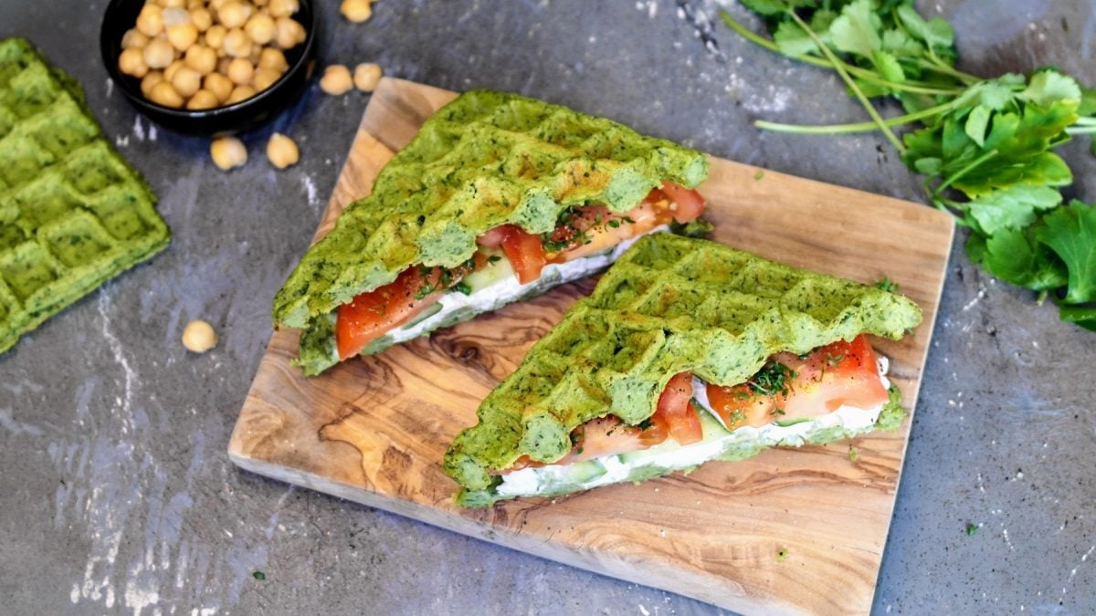 Ein kleines Holzbrett mit dem Falafel-Waffel-Sandwich auf einem steinernen Tisch. Ebenso oben im Bild sind frische Kräuter und Kichererbsen. Alles von schräg oben fotografiert.